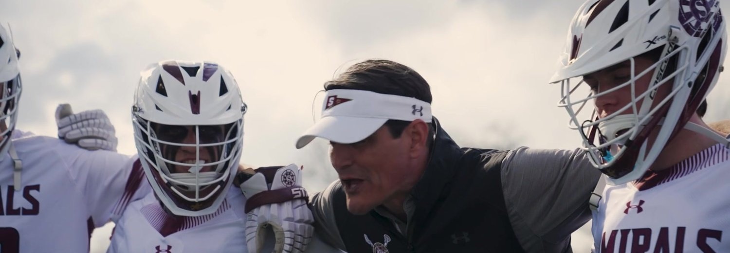 Coach in a visor talks with helmeted high school lacrosse players in a sideline huddle