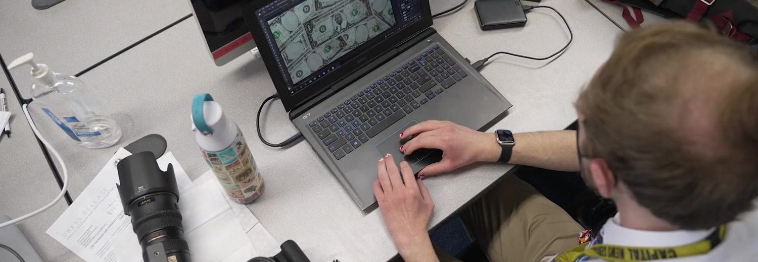 Over-the-shoulder view of a student editing video on a laptop beside cameras and lenses
